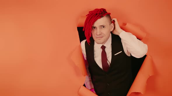 Thoughtful Young Man Scratching Back of Head and Looking Out of Hole of Orange Background