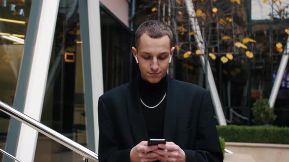 Young Businessman Using His Smartphone and Smiles. He Is Texting Messages While Standing Near Modern