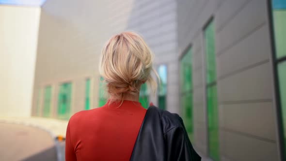 a Blonde in a Red Dress with a Black Jacket Over Her Shoulder Walks Along the Wall with Large