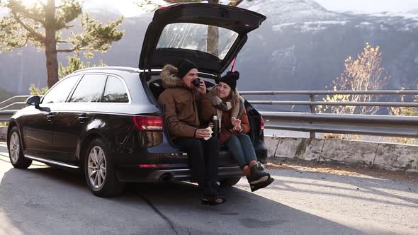 Loving Young Couple Drinks Hot Tea From Thermos Flask Sitting in Car Trunk