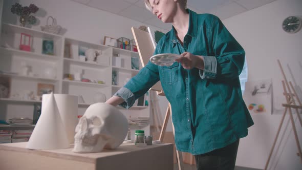 A Young Woman Artist Painting in the Art Studio - a Skull and Other Figures on the Table