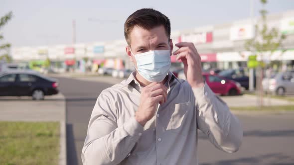 A Young Caucasian Man Puts on a Face Mask and Looks at the Camera in an Urban Area