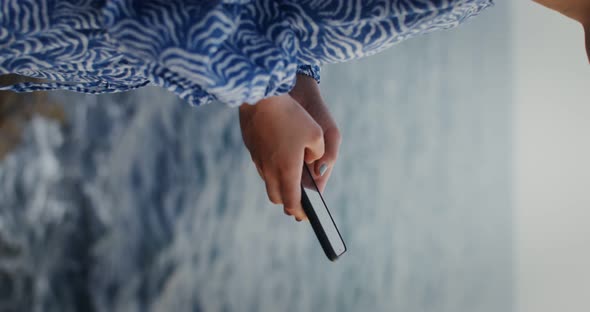 A Woman is Typing on a Mobile Phone While Standing on the Rocks on the Seashore