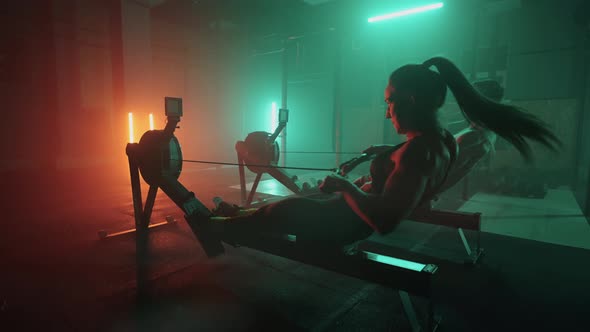 A Man and a Woman in a Colored Neon Red and Blue Light Do a Workout on a Rowing Simulator Together