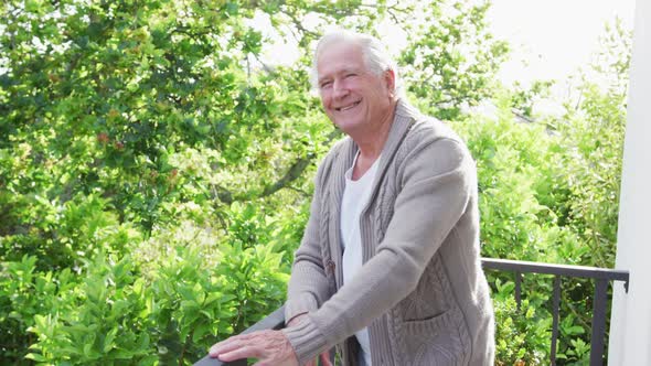 Portrait of smiling retired senior man wearing cardigan sweater leaning on railing in balcony