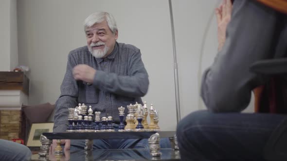 Portrait of Embarrassed Old Caucasian Man Sitting in Front of Chess Desk and Smiling. Senior Retiree