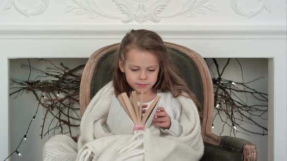 Lovely Little Girl Wrapped in White Christmas Blanket Exploring a Book Sitting in an Armchair Near