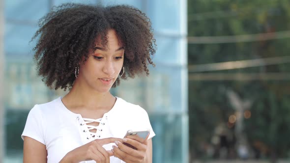 Smiling Young Afro American Woman with Interest Looks Into Smartphone, Excitedly Happily Points