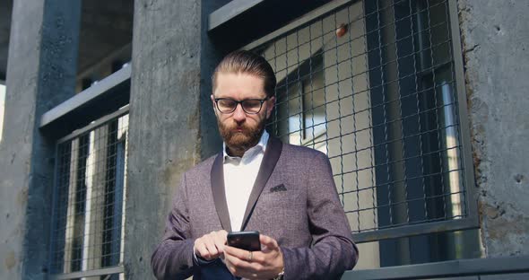 Man in Glasses which Waiting for Somebody while Using his Phone near wall of modern building
