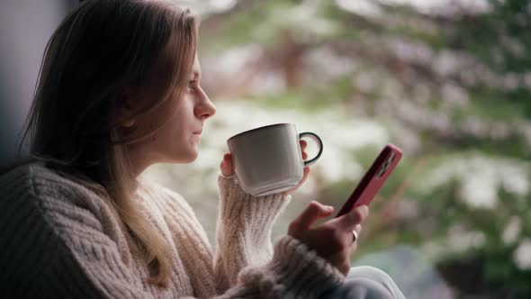 A Young Girl Sits on the Windowsill and Drinks Coffee While Using Her Smartphone