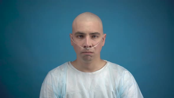 A Bald Young Man with Oncology Sadly Looks at the Camera on a Blue Background. Hair Loss Due To