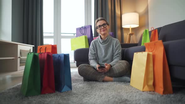 Portrait of a Happy Woman Sitting on a Carpet in a Cozy Room Among Shopping Bags