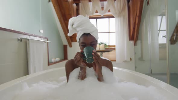 African american attractive woman relaxing in foam bath and drinking coffee in bathroom