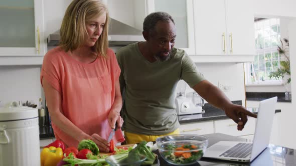 Diverse senior couple preparing food in kitchen following recipe on laptop