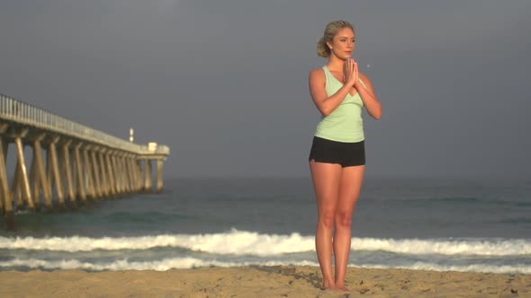 A young woman does yoga on the beach next to a pier.