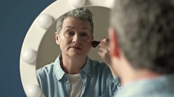 Wellgroomed Elderly Woman Applies Blush on Her Cheek with a Makeup Brush Sitting Alone at Home in