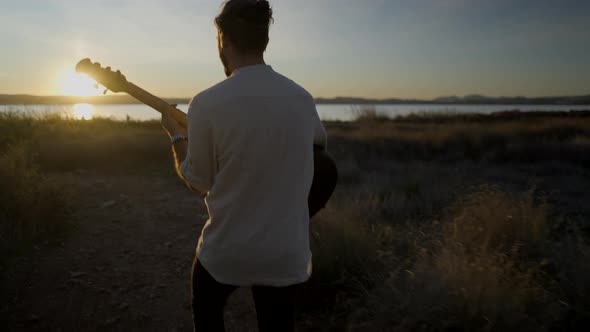 Guy Holding Guitar and Walking Towards the Lake to See the Picturesque Sunset