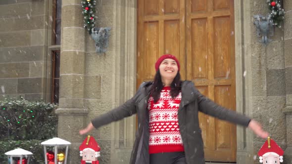 A Happy Young Woman in a Sweater with Reindeer Comes Out of the House Into the Street