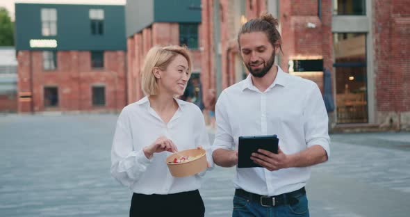 Business Partners Caucasian Man and Woman Walking in the Evening City Eating Salad and Using laptop.