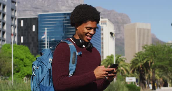 Smiling african american man using smartphone, wearing headphones and backpack in city street