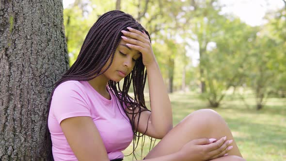 A Young Black Woman Thinks About Something As She Sits Under a Tree in a Park on a Sunny Day