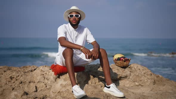 Wide Shot of Thoughtful Calm African American Young Man Sitting in Sunshine on Cliff with Fruits As