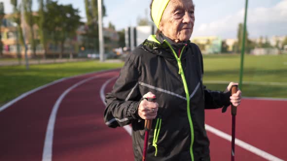 Senior Woman Walking with Walking Poles in Stadium on a Red Rubber Cover. Elderly Woman 88 Years Old