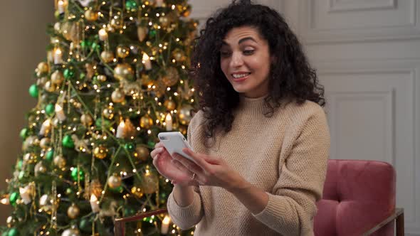 Young Woman with Curly Hair Using a Smartphone Sitting on the Sofa By the Christmas Tree Celebrating