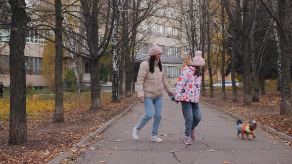 Mom and Daughter are Walking Along an Autumn Alley with a Dog