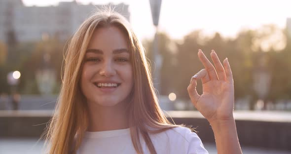 Outdoors Portrait of Attractive Young Millennial Girl Smiles at Camera Showing Hand Ok Sign