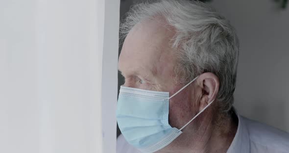 Worried Senior Man in Medical Mask Looking Through Window, Leaning To Wall