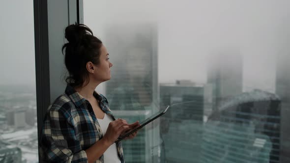 Attractive Girl Stands at the Window Against the Background of the City of Skyscrapers and Works