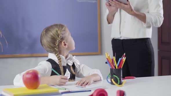 Smart Caucasian Girl with Pigtails Looking Back at Unrecognizable Tutor and Writing in Exercise Book