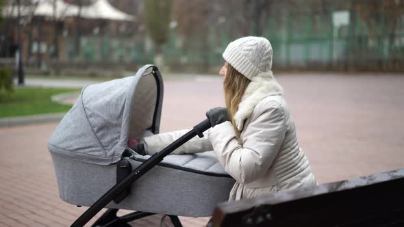 Young Mother with Newborn Child Outdoor. She Sitting on the Bench with Baby Sleeping in Pram