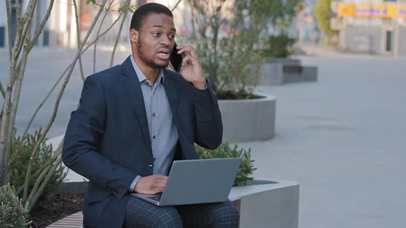 Focused African Businessman Holding Smartphone Look at Laptop Screen Make Business Call Notes