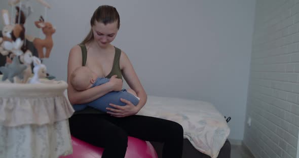 Happy Young Mother Lulls and Breastfeeds Her Little Baby on Fitball in Bedroom