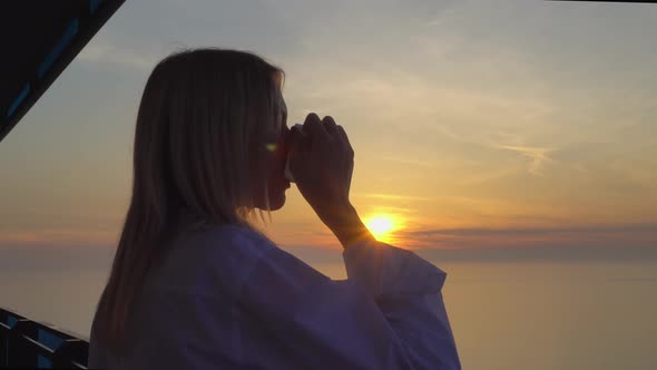 a woman admires the sunset over the sea, and drinks coffee on a high floor.