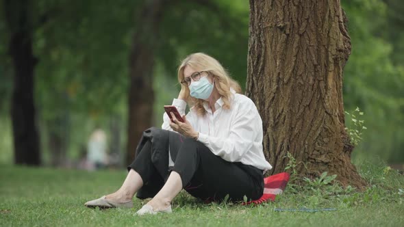 Wide Shot of Relaxed Middle Aged Caucasian Woman in Covid19 Face Mask Sitting at Tree in Park