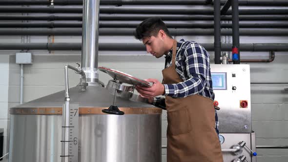 Handsome Brewer in Uniform Working at the Brewery with Metal Containers on the Background