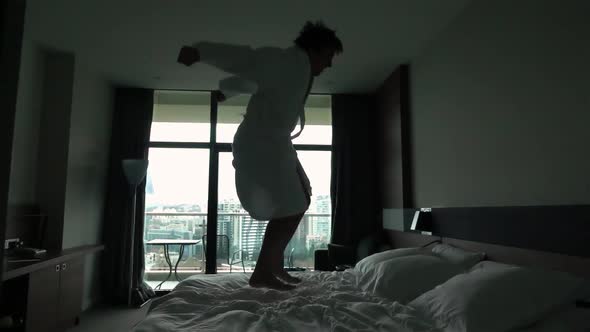 Happy Young Man Jumping on Bed at Hotel in Bathrobe