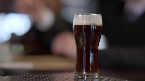 Closeup Glass with Dark Lager Beer on Bar Counter with Blurred Male Clients at Background Drinking