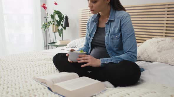 Caucasian woman in advanced pregnancy sitting on bed, eating salad and reading book. Shot with RED h