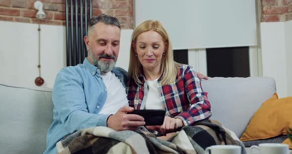 Man and Woman Resting on Sofa with Blanket on their Legs and Reviewing their Photos on Mobile