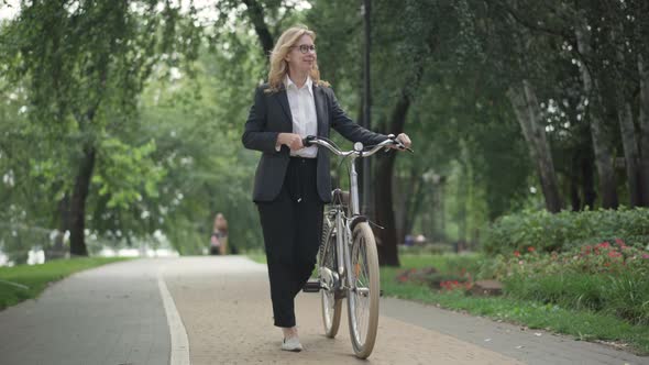 Wide Shot Portrait of Smiling Successful Caucasian Businesswoman Walking with Bike on Summer Park