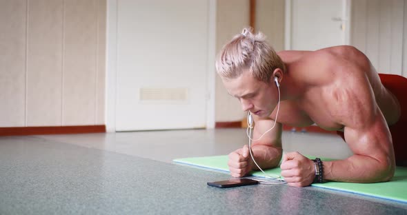 A Young Muscle Shirtless Man Does Plank Exercises and Listens to Music Through an Online Smartphone