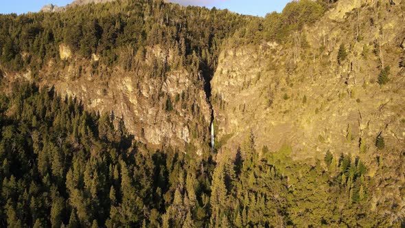 Aerial dolly out over Corbata Blanca waterfall hiding in mountains surrounded by woods at golden hou