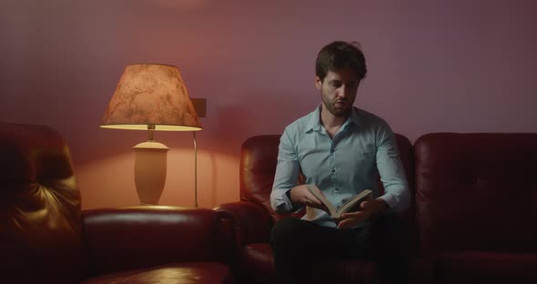 Young guy with shirt reads a book sitting in the living room at home