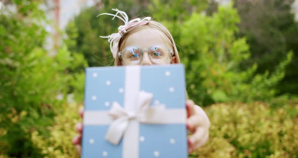 A Beautiful 7Yearold Girl with Glasses Give a Gift in a Blue Box with a Bow