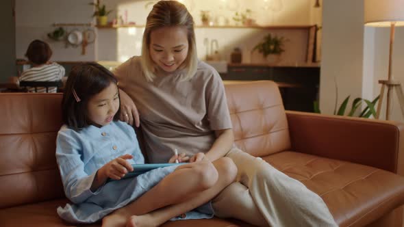 Loving Asian Mom and Little Daughter Sit Together on Sofa at Home With Tablet Device Draw With