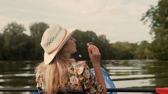 Girl Enjoying At Sunset. Blonde Woman Blowing Soap Bubbles. Woman In Hat Relaxing On Kayak Boat.